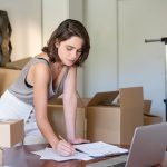 Empreendedora administra seu negócio de roupas em um escritório doméstico, diante de um laptop e de caixas para envio.