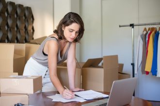 Empreendedora administra seu negócio de roupas em um escritório doméstico, diante de um laptop e de caixas para envio.